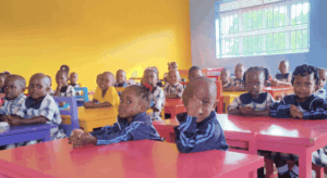 kinder-klassenraum-vorschule-buwooya-uganda Kinder sitzen an bunten Tischen in einem Klassenraum der Fransalian Vorschule in Buwooya.
